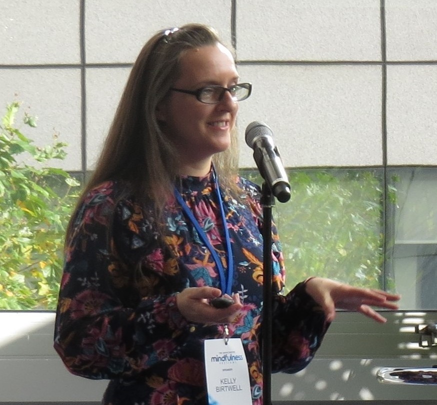 Doctor Kelly Birtwell, a white woman with long brown hair and dark-rimmed glasses, wearing a dark blue and pink patterened dress.
